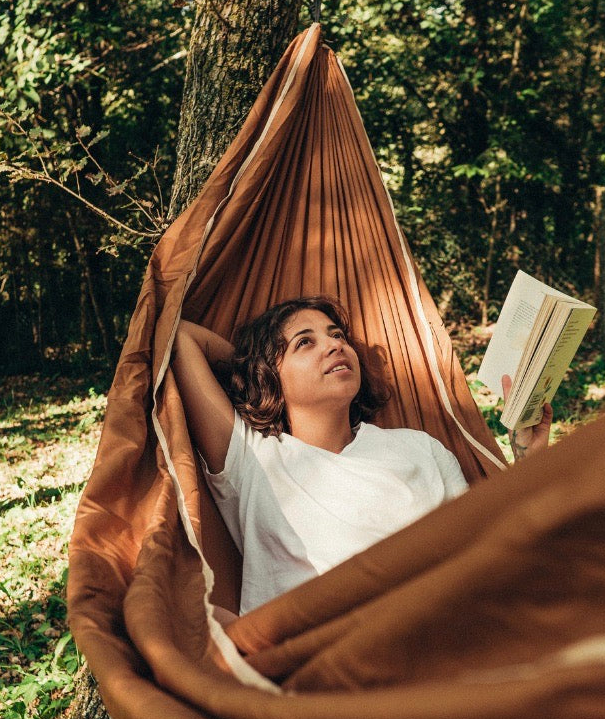A teen girl relaxes in a brown fabric hammock strung between trees in a wooded outdoor setting, holding an open book.