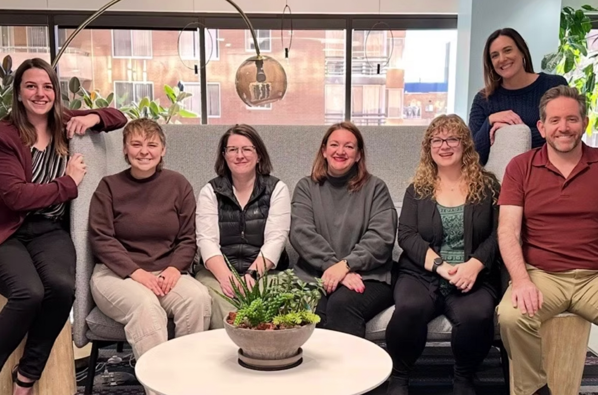 Seven people posing together in a modern office lobby setting, seated on and around a gray sofa with a white coffee table in front featuring a potted succulent.