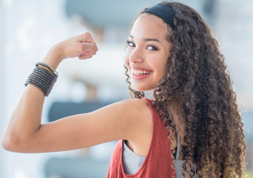 A smiling girl with long curly brown hair flexes her bicep, wearing a rust-red tank top, gray undershirt, black headband, and a black beaded bracelet.