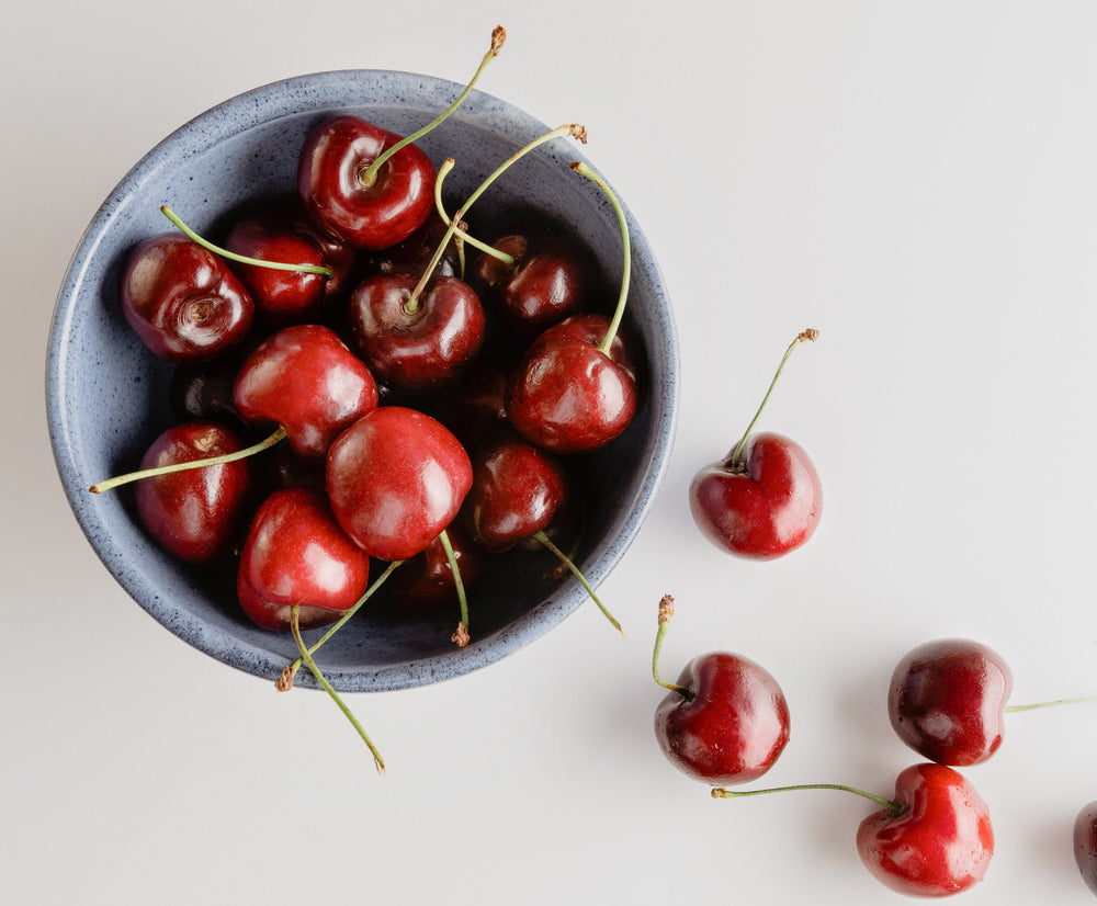 A top-down view of a speckled blue-gray ceramic bowl filled with glossy red cherries, with a few cherries scattered on a clean white surface.